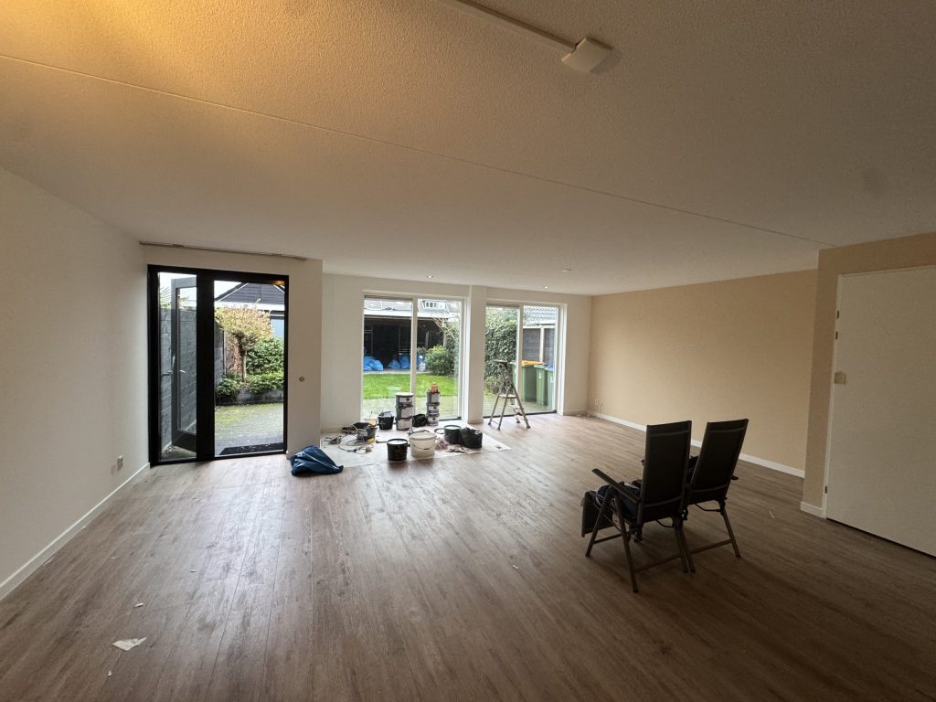 Lege woonkamer van een bestaande rijwoning met laminaat, tuindeuren en een ladder bij de schuifpui